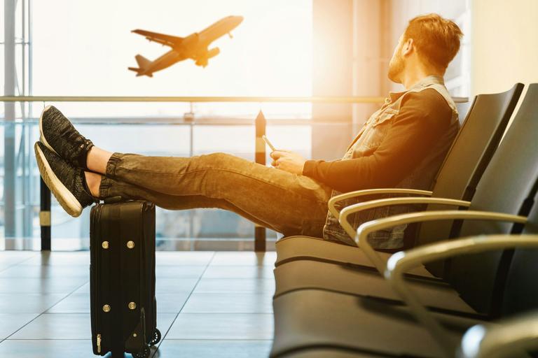 Mann sitzt entspannt in Flughafen-Lounge, legt Füße auf Trolley-Koffer und blickt auf startendes Flugzeug durch Panoramafenster.